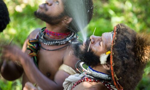 Two men in traditional clothing are spraying water on their faces.
